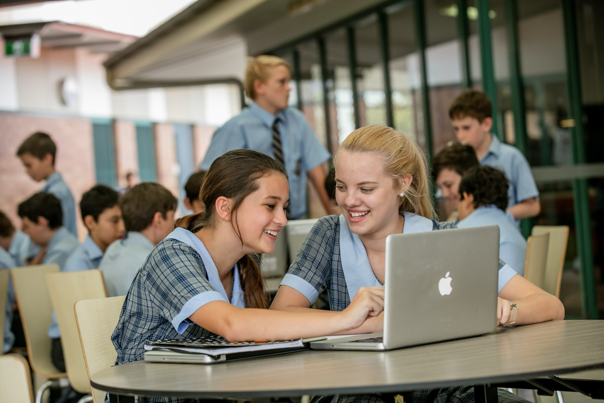 Two students in school uniforms sitting at a table working on a laptop
