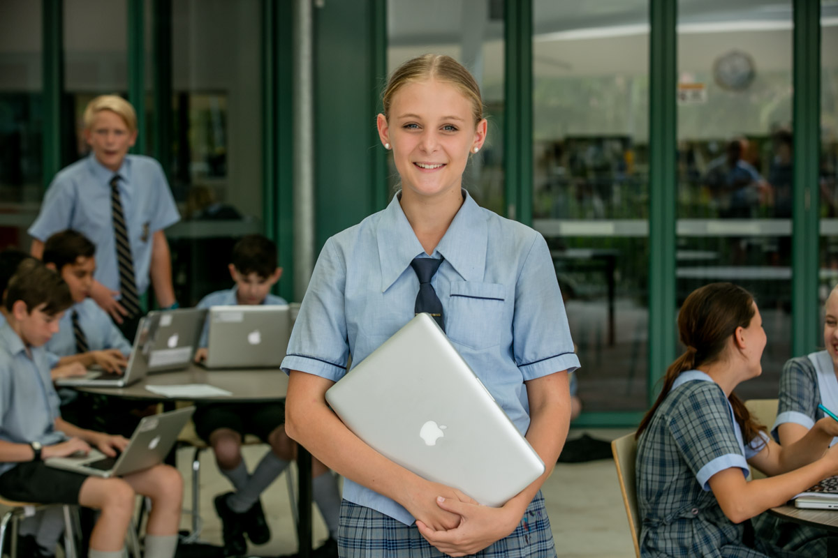 Student in school uniform holding a laptop while other students work on laptops in the background