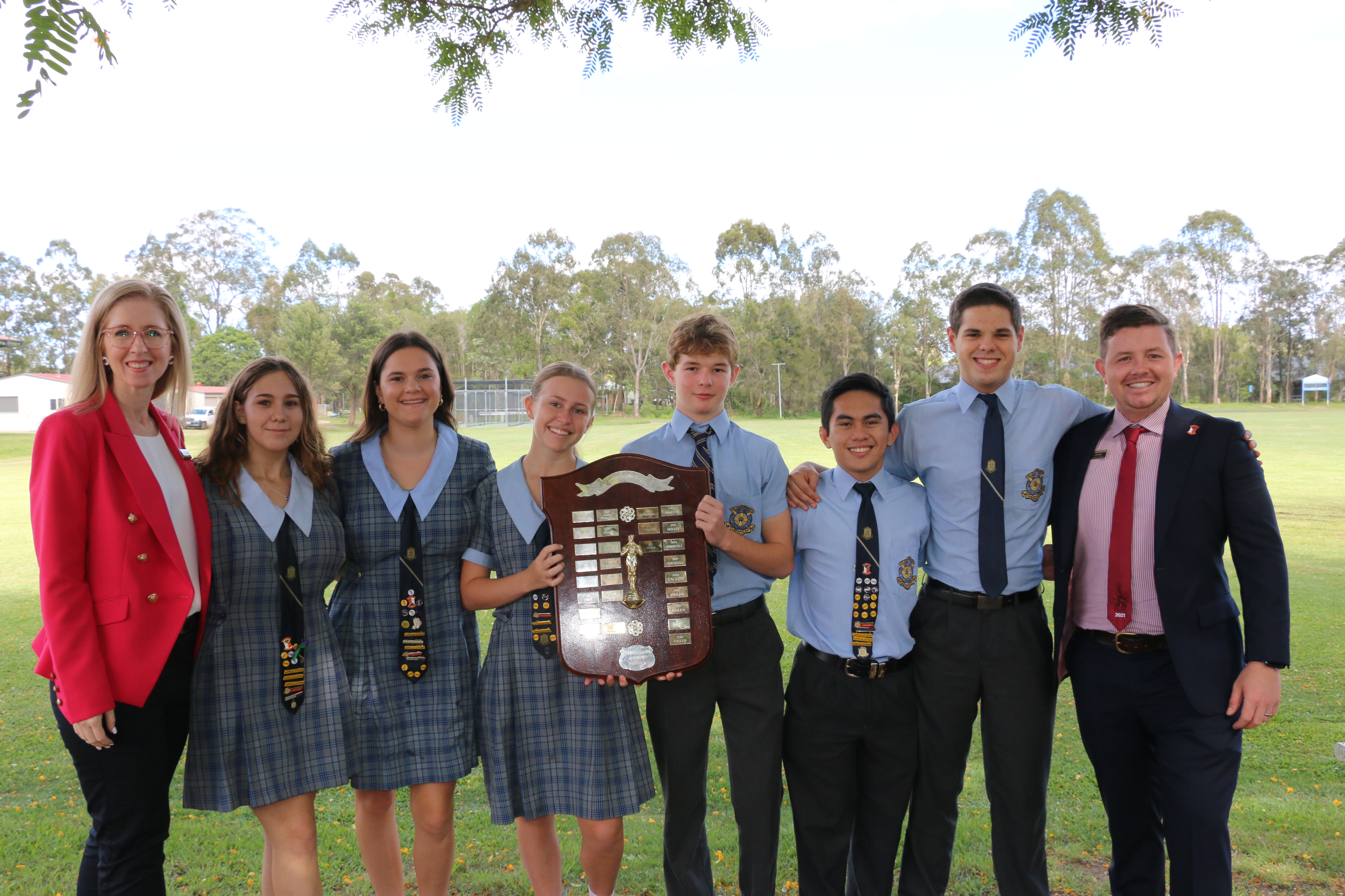 Group of students and two adults standing outdoors holding a large shield‑style award