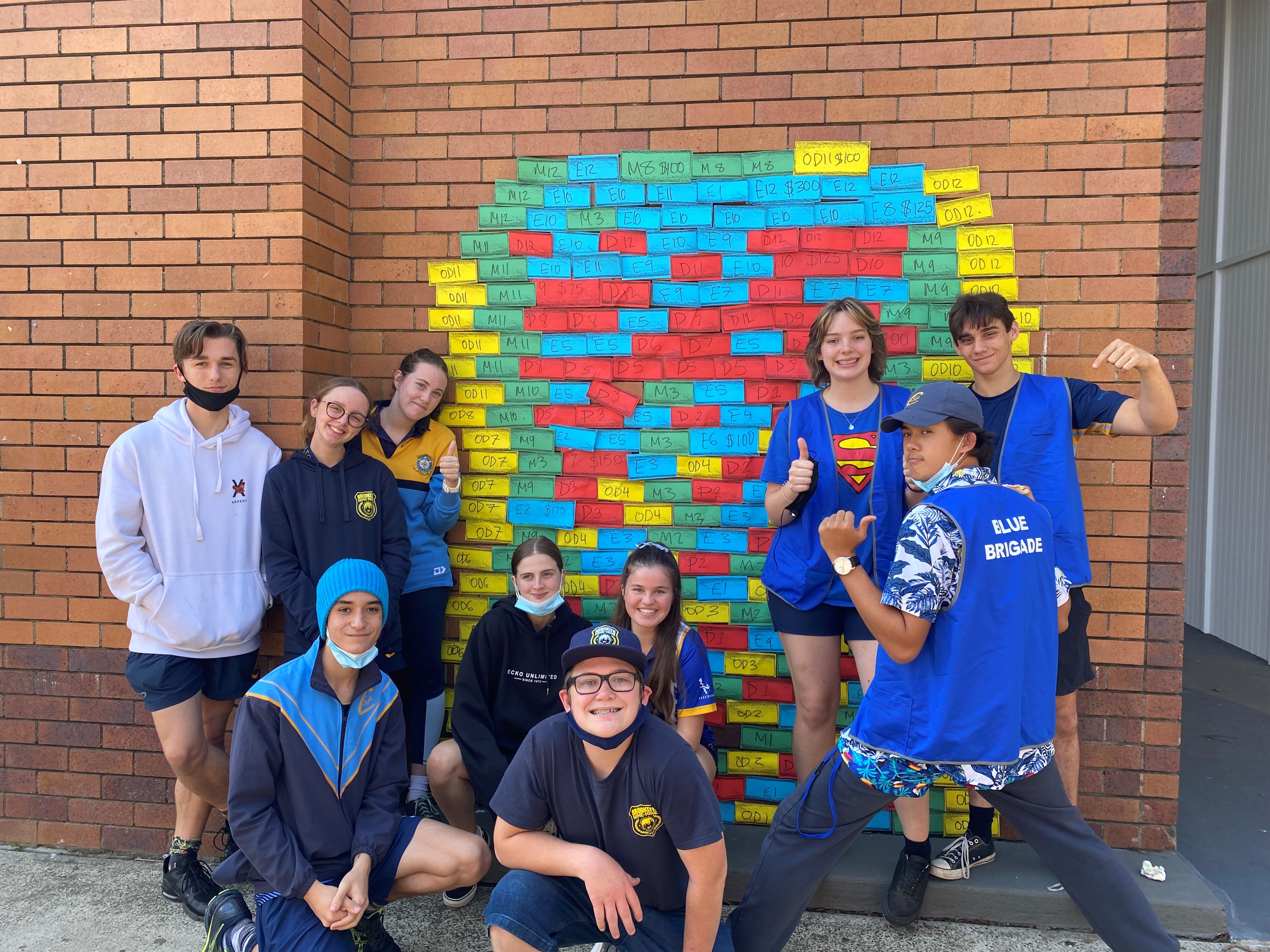 Group of students standing and kneeling in front of a colourful brick-style display on a brick wall