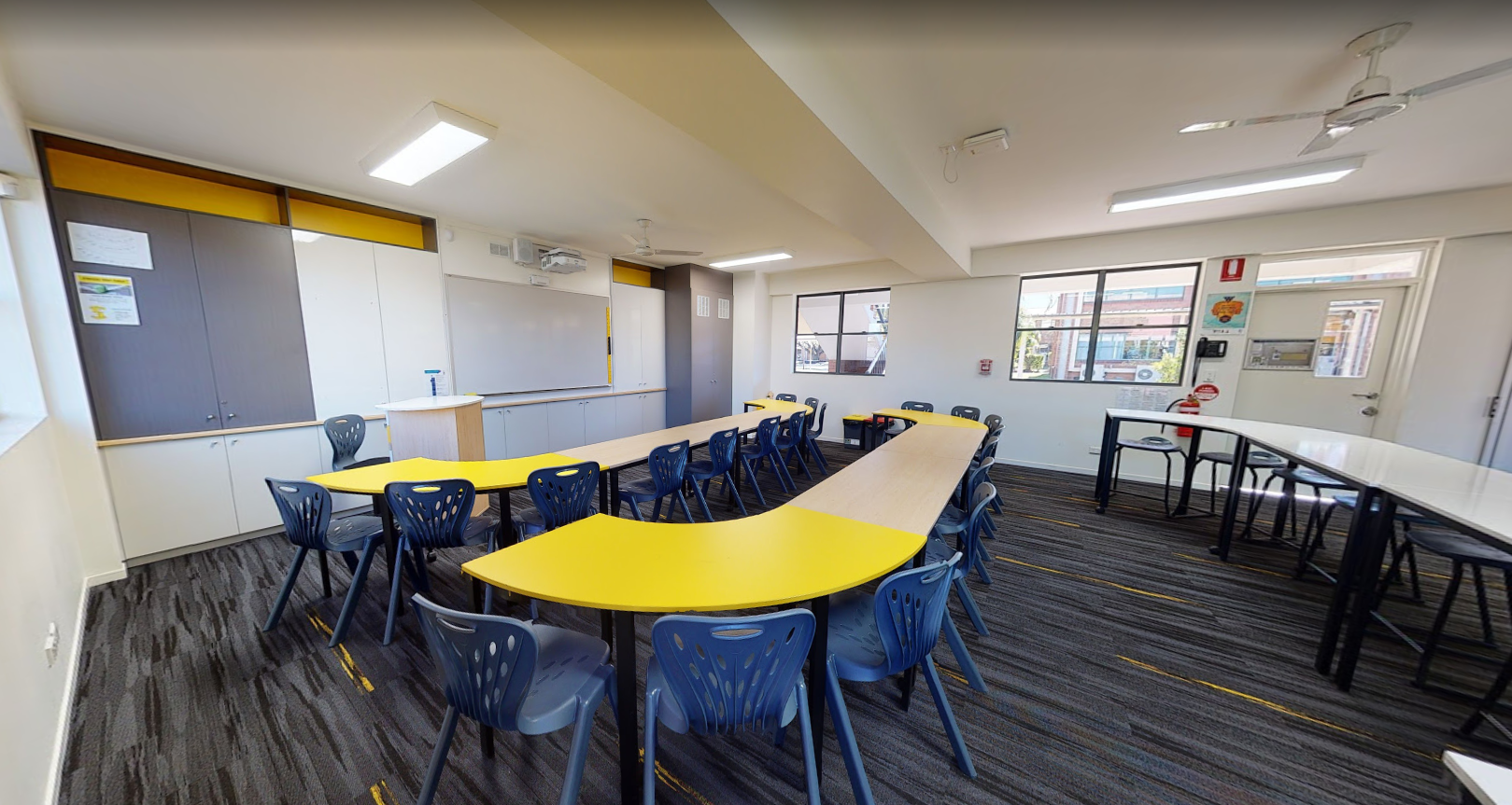 Modern classroom with curved yellow desks, multiple chairs, whiteboard and large windows providing natural light