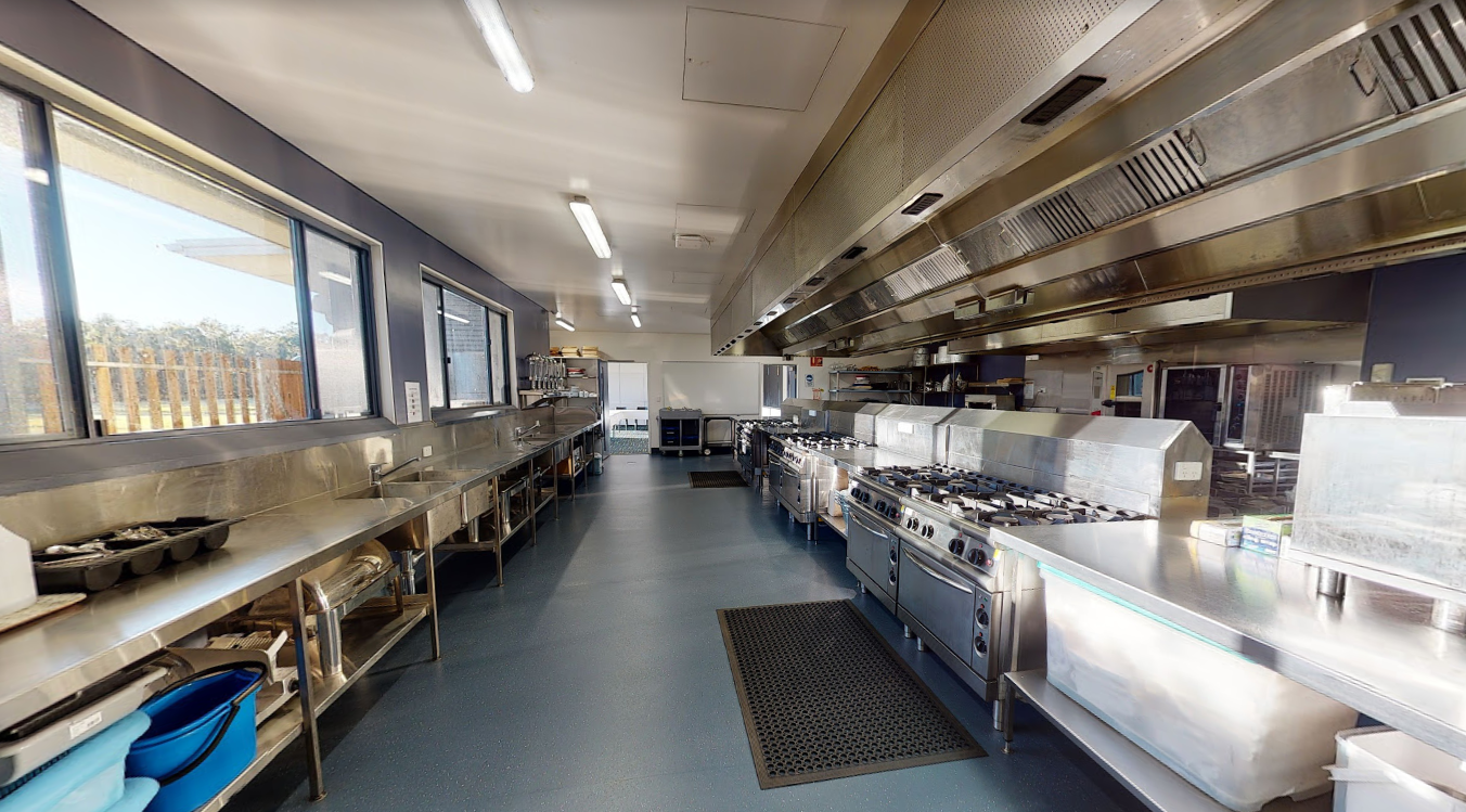 Large stainless‑steel commercial kitchen with multiple ovens, stovetops, benches and overhead ventilation