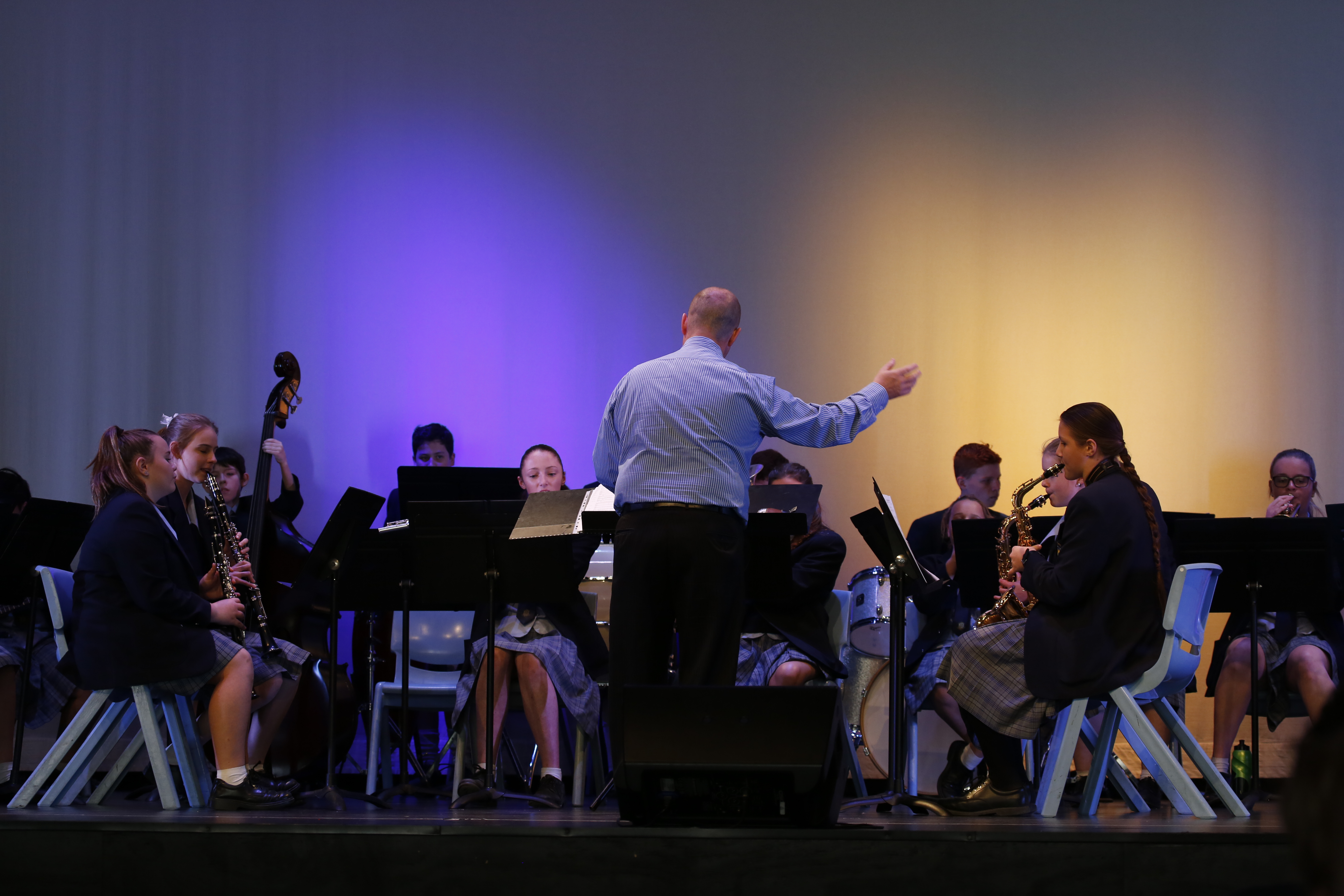 Conductor leading a seated student ensemble on stage under coloured lighting