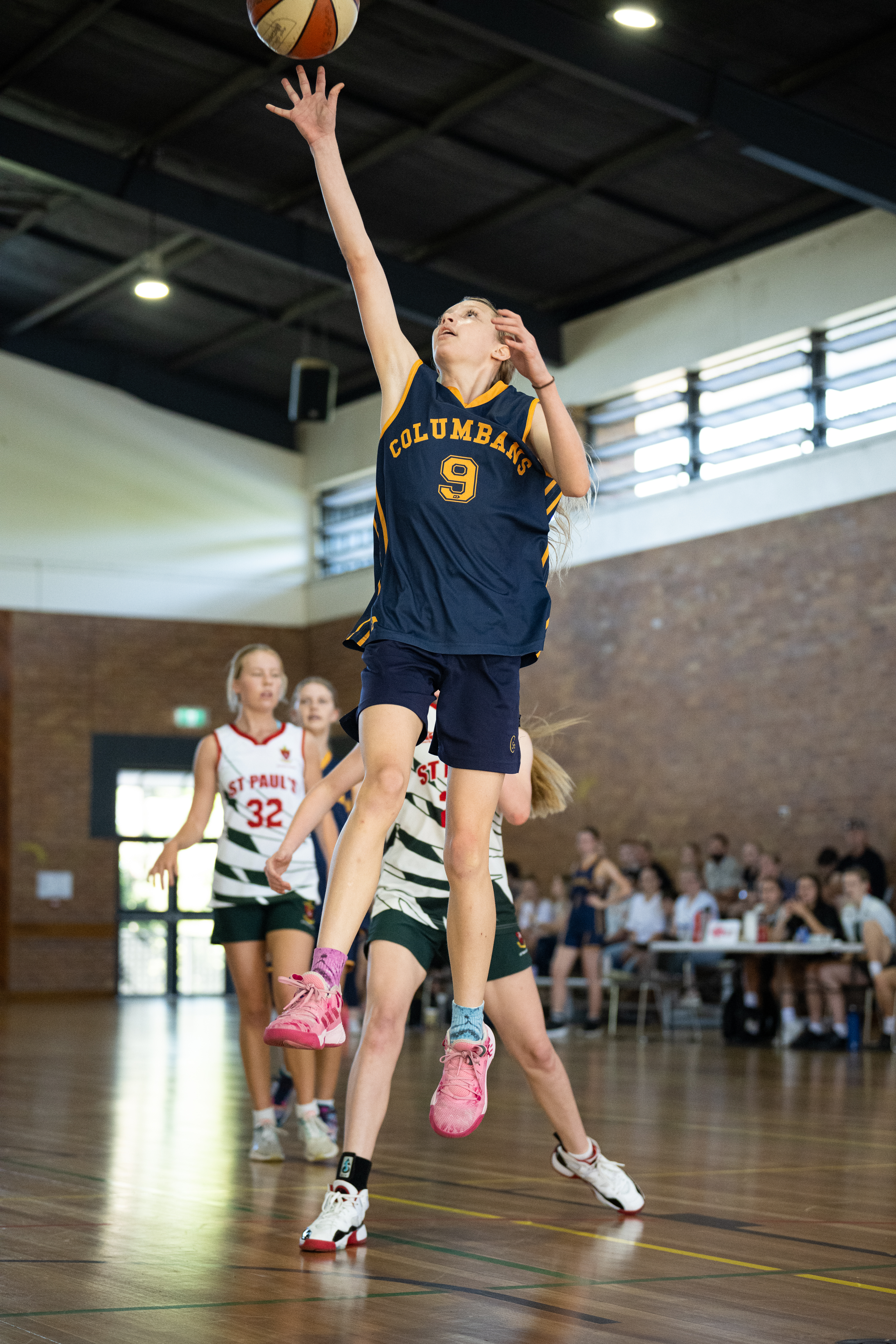 Basketball player in a Columbans jersey jumping to shoot the ball during an indoor game