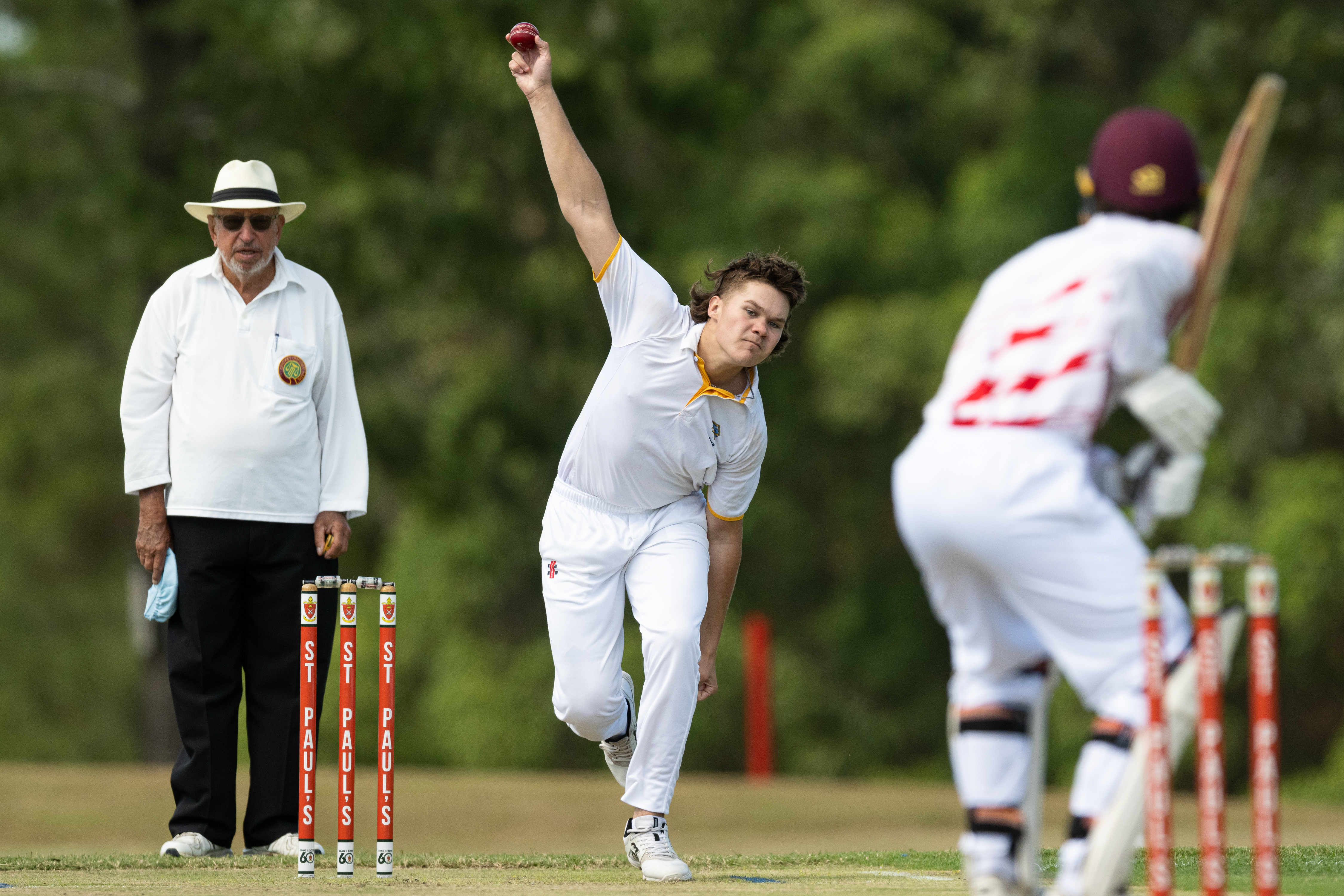 Bowler delivering a cricket ball toward a batter with an umpire standing behind the stumps