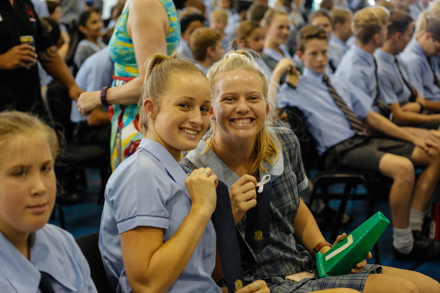 Two students in school uniforms sitting in an assembly holding up ties.
