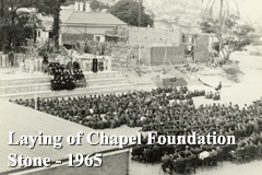 Historic photo of a large gathering seated outdoors with the text ‘Laying of Chapel Foundation Stone – 1965