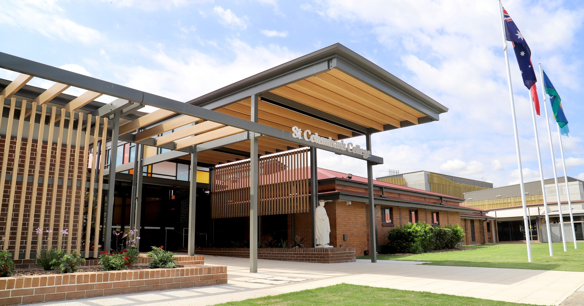Front exterior of St Columban’s College with modern entry structure, brick buildings, landscaped gardens and three flagpoles
