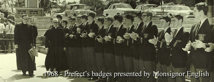 Historic group photo showing students in uniform standing in a row while two clergy members stand nearby, with the text ‘1968 – Prefect’s badges presented by Monsignor English’ displayed at the bottom
